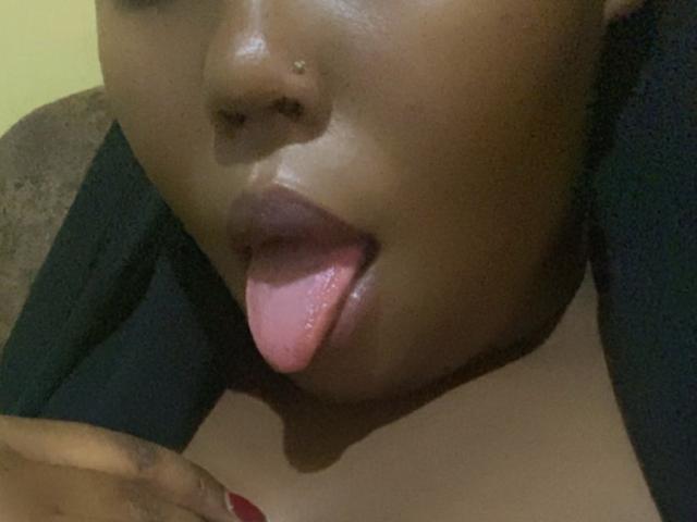AnitaNini's dark brown face with shiny skin, nose piercing, and pink tongue sticking out, showing red nails on the left side. Her black clothes are partially visible.