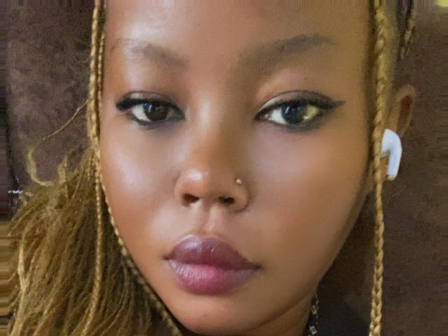 Close-up of AnitaNini's face. She has dark brown skin and wears blonde braided hair on both sides. Her eyes are almond-shaped with black eyeliner, and she has a small silver nose stud on her right nostril. She wears white earphones in both ears. Her lips are full and painted a dark pink. The texture of her skin is smooth. The image is slightly blurry. The background is out of focus, showing a brown, textured surface.