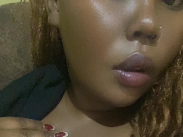 Close-up photo of AnitaNini's face. Her dark brown skin is glossy, with visible moisture on her cheeks and lips. She has full, slightly parted pinkish-gray lips. Her curly, light brown hair frames the upper part of the image. She's wearing a black garment, with a hand with red and white nail polish touching her collarbone. The background is blurred and indistinct. The focus is on her face and lips, highlighting her natural beauty and glossy skin texture.