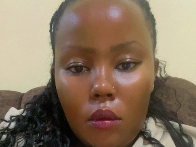 Close-up of AnitaNini with dark, shiny skin and defined, symmetrical facial features. Her black, curly hair is pulled back, and she has a small nose stud in her left nostril. She wears black eyeliner with a slight wing, and her lips are full with a glossy pinkish-red color. Her expression is neutral, slightly downward gaze. She's wearing a beige top. The lighting highlights her smooth skin and subtle sheen. The background is a plain, light-colored wall. The image is slightly blurry.