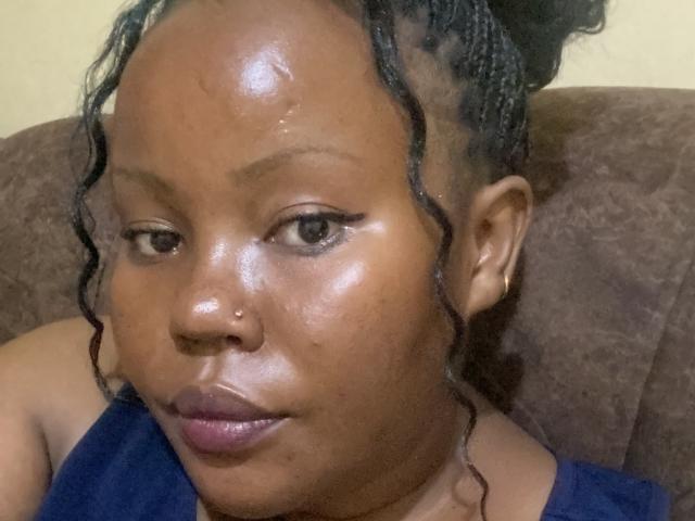 Close-up photo of AnitaNini with medium-dark brown skin and a shiny, slightly oily complexion. She has a small, black nose ring in her right nostril and dark, slightly curled hair pulled back into a small bun. Her eyes are brown and she has thin, black eyebrows. She wears a dark blue sleeveless top. Her lips are full and have a natural pinkish tint. The texture of her skin is smooth with visible sheen. The background is a brown, textured fabric. Her expression is neutral, with slightly parted lips.