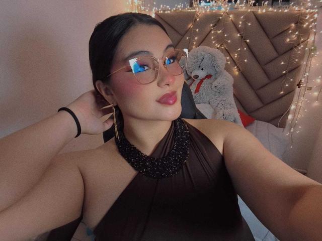 HelueJamil with glasses, pink blush, shiny pink lips, black necklace, brown dress, sitting, black chair, dark hair in a ponytail, smiling, cute, curvy, small earrings, soft lighting.