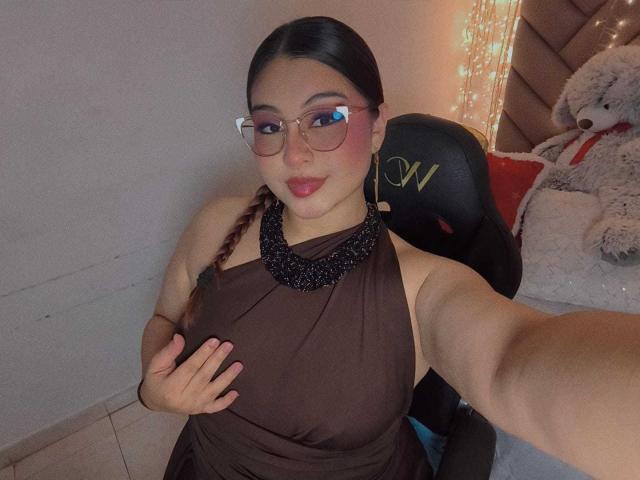 HelueJamil with glasses, black sheer dress, black bracelet, and pink blush, posing with finger on chin, showing tits, and a cute smile, next to a stuffed teddy bear.