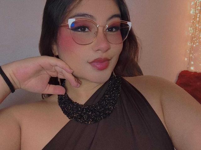 HelueJamil with puffy pink lips, blue eyes, and glasses, wearing a black halter top, black bracelet, and gold earrings, has a white fluffy dog with a red bow.
