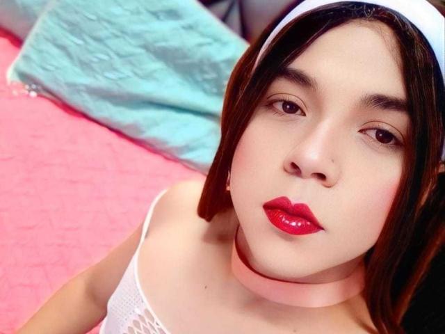 AndrewTaylor69 with short brown hair, white headband, pink choker, red lipstick, white lace top, pink carpet, and blue pillow, looking up at the camera with a neutral expression.