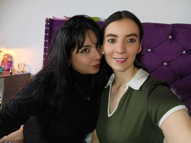 Two ValkiriaAndDiones with black hair and green eyes, one in a black top and the other in a green shirt with white collar, smiling and standing next to a purple headboard.