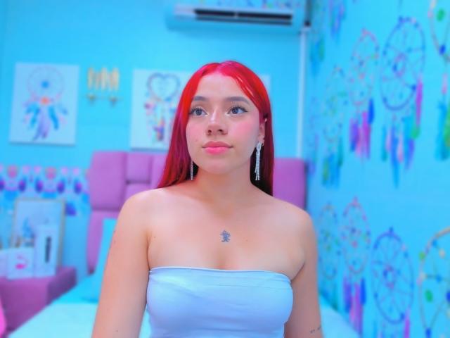 MiaLeroy, con cabello rojo, vestida con un top blanco sin tirantes que deja ver sus pechos, tiene un pequeño tatuaje en el pecho y está de pie en una habitación azul, como en un sueño..