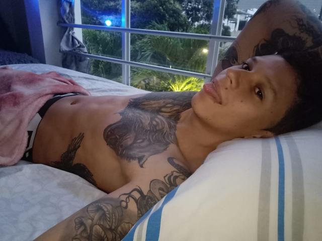 MasterDaveExtreme lying on a bed, shirtless, with tattoos on chest and arms, puffy lips, short curly hair, wearing black underwear, pink blanket, and white striped pillow.