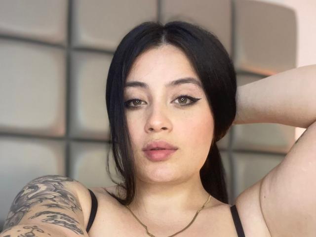 Close-up of AnnyBentleii, a young woman with long black hair, light skin, and tattoos on her right arm. She has full lips, green eyes, and wears a black strap top. Her hand rests behind her head, creating a sexy, confident pose.