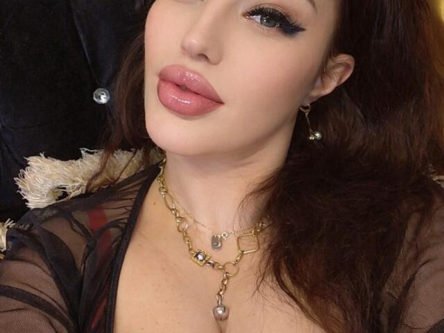 Vanessa, con labbra rosa lucide, eyeliner nero e capelli castano ondulati, indossa una collana dorata con una perla, e una maglia nera trasparente. Il suo seno è leggermente visibile..