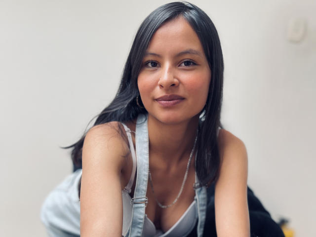 Foto van LillyBonnet, een aantrekkelijke Aziatische vrouw met lang zwart haar, die een witte tanktop en een zilveren ketting draagt. Naar voren leunend, licht glimlachend, met zichtbaar decolleté en zacht licht.