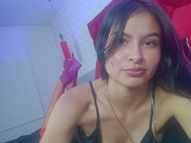 LillyBonnet ha i capelli neri, occhi marroni, e un leggero sorriso. Lei sta tenendo un dildo rosso nella mano destra, indossando una canotta nera con spalline sottili e una collana d'oro.