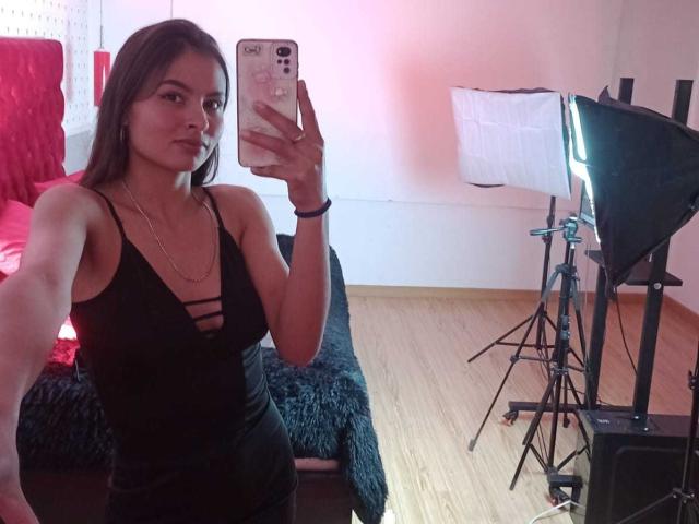 LillyBonnet prend un selfie en miroir dans une pièce bien éclairée. Elle porte une robe noire à bretelles avec un décolleté profond, tenant un téléphone dans sa main droite. Installation d'éclairage sur la droite.