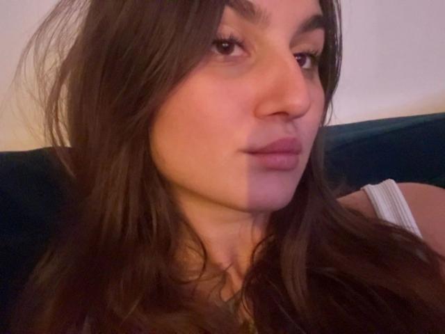 Close-up selfie of MilaSusse-hot, showing her face and upper shoulders. She has long, dark brown hair, slightly parted lips, and a subtle, suggestive expression. Dim lighting creates a soft glow.