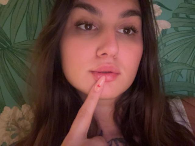 Nahaufnahme von MilaSusse-heiß, mit langen braunen Haaren, braunen Augen und einem Finger, der ihre leicht geteilten Lippen berührt. Sie hat einen Nasenring und ein Tattoo auf der Brust. Teal grünen Hintergrund.