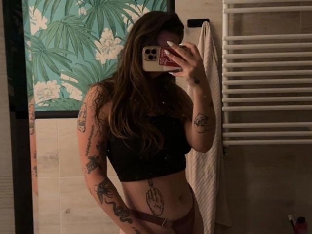 Selfie de MilaSusse-hot, mulher tatuada com cabelos castanhos longos, top preto, segurando o telefone, cobrindo o rosto, mão visível tatuagens, parede com padrões florais, radiador de toalha no fundo, iluminação fraca.