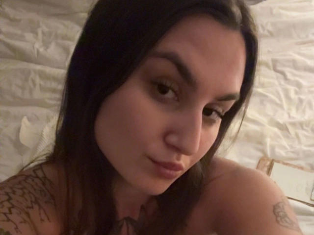 Close-up foto van MilaSusse, een aantrekkelijke, donkerharige vrouw met tatoeages op haar schouder, die naar beneden kijkt met een subtiele glimlach. Zacht, gedimd licht, blote schouders, subtiele schaduwen, suggestieve blik.