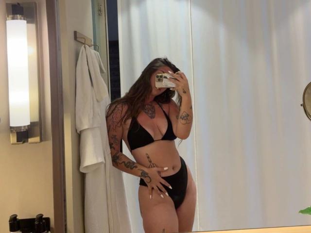 Spiegel selfie van MilaSusse-hot in een zwarte bikini, pronken met tatoeages op haar armen en zijde. Lang bruin haar, telefoon in linkerhand, pik zichtbaar onder bikinibroekje. Een lichte badkamer.