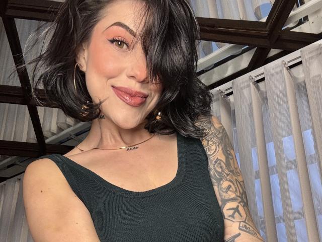 Fotografia de HannaBorbon com cabelo preto na altura dos ombros, sorrindo com batom vermelho, vestindo uma blusa preta sem mangas. grandes brincos de argola, e um braço tatuado. A luz filtra através das persianas atrás dela.