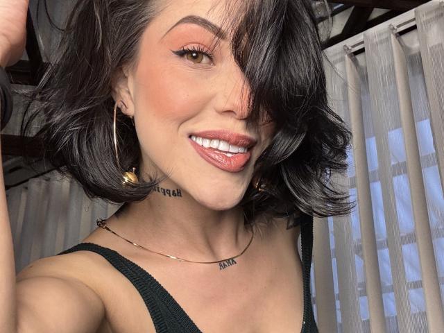 Primer plano de HannaBorbon, sonriendo con labios rosados brillantes, pelo negro hasta los hombros, vistiendo una camiseta sin mangas negra, aro dorado pendientes, y un delicado collar. Fondo: persianas verticales, luz del día brillante. Mierda de caliente.