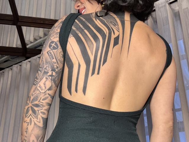 Fotografie von HannaBorbon mit dunklen, welligen Haaren, Rücken zur Kamera, schwarzes Tanktop, das ein großes, schwarzes Stammes-Tattoo zeigt. Auf ihrem Rücken und detaillierte Arm Tattoo. Ficken sexy, tätowiert, ausgesetzt zurück.