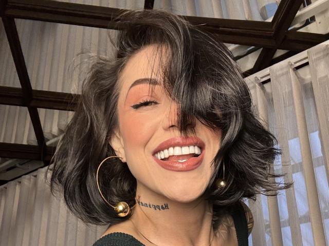 Primer plano de HannaBorbon, sonriendo con la lengua hacia fuera, pelo ondulado negro que cubre un ojo, grandes pendientes de aro de oro, "respirar" tatuaje en el cuello, techo de madera y cortinas grises en el fondo. Mierda sexy.