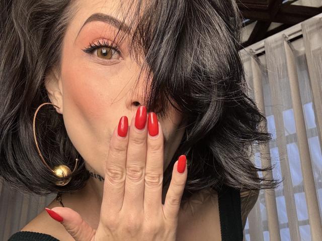 Gros plan de HannaBorbon avec des cheveux foncés et ondulés couvrant un œil, des ongles rouges, une main partiellement sur sa bouche, un grand cerceau boucle d'oreille avec boule dorée, dessus noir, sexy, regard intense, fard à paupières rouge.