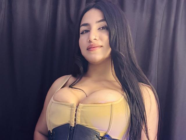 AlanaHarris with long black hair, tan and black corset, large tits, slight smile, standing against dark curtain, looking at camera, soft lighting, slight pink lipstick.