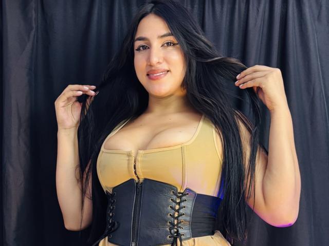 AlanaHarris has long black hair, wearing a yellow top with a black lace-up corset, showcasing her big tits. She's smiling, holding her hair, against a black curtain.