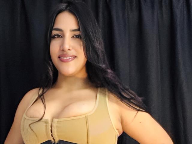 AlanaHarris with long black hair, tan tank top showing her big tits, smiling at the camera, dark curtains in the background. She has light skin and brown eyes.