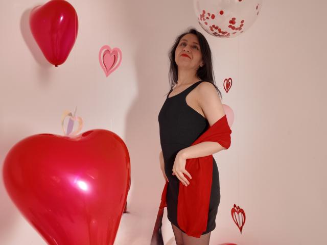 Cyntiia with black tank dress, red jacket, and pantyhose, standing with red balloons and hearts, looking up, hands on hip, long dark hair, fair skin, slight smile.