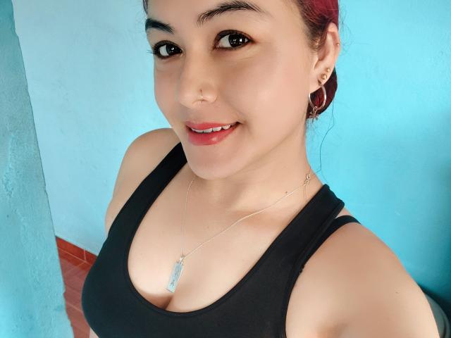 Thalassa with red hair, black tank top, cute nose ring, hoop earrings, silver necklace with blue stone, smiling at camera, showing her tits and cute face.