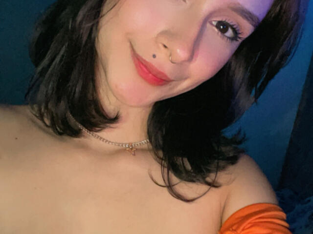 CamilaJolie has light skin, dark brown hair, a nose ring, and a small mole under her right eye. She's smiling with red lips, wearing an orange off-shoulder top.