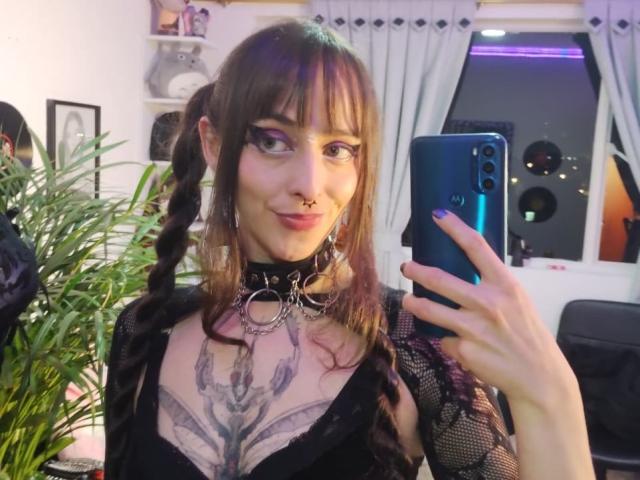 KizzuKyuwu with braided brown hair, black lace top, collar with metal rings, nose ring, and large chest tattoo, holding a blue phone, smiling.