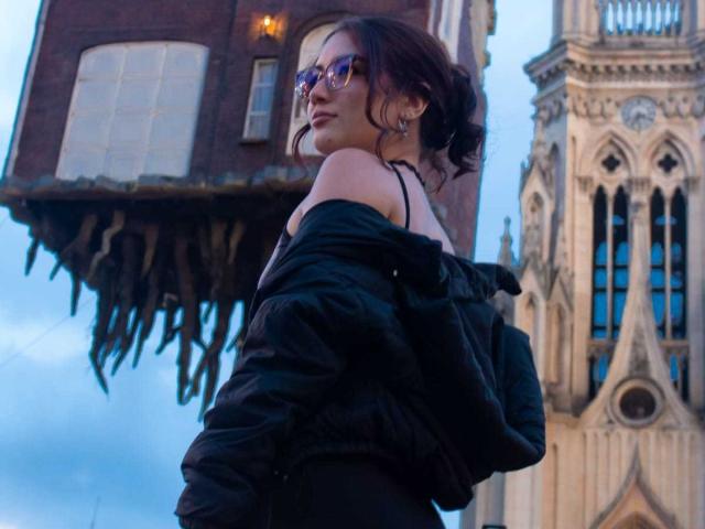 Stardustt with glasses, black off-shoulder top, and black jacket, looking over her shoulder, standing in front of a floating brick house and Gothic-style tower.
