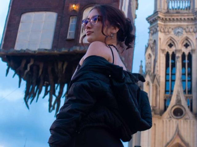 Stardustt with red hair, glasses, and black off-shoulder jacket, looking back, standing in front of a floating brick house and Gothic tower. Her ass is slightly visible.