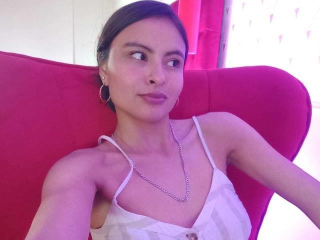 LillyBonnet with short brown hair, wearing a white patterned top with thin straps, small tits, silver chain necklace, and hoop earrings, sitting on a red chair, pouting.