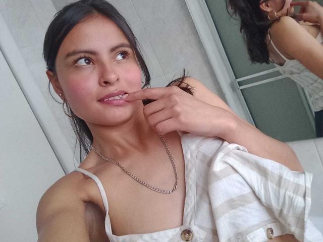 LillyBonnet, a young woman with medium brown skin and black hair, is in a selfie. She has her finger lightly touching her cheek, smiling slightly. She wears a white, striped, button-down top with thin straps and a silver chain necklace. Her reflection is visible in a mirror on the right. She has small earrings and her hair is pulled back. The photo is clear and focused. Her expression is relaxed and slightly playful.