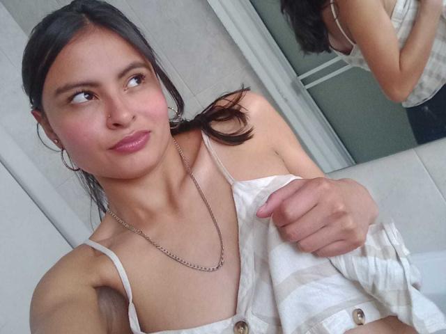 LillyBonnet has a cute smile, pink lips, and is wearing a white striped top with a chain necklace. She's holding her top, showing small, perky tits. Reflection in mirror.