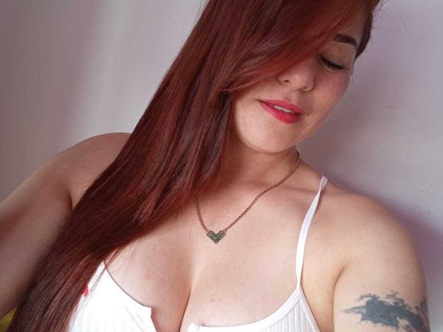 Casssandra with long red hair, white tank top, and green necklace, showing her tits, smiling, and tattoo on right arm. She's wearing pink lipstick and a gold necklace.