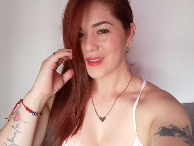 Cassandra has red hair, white top, small green pendant, tattoos on arms, red bracelet, gold hoop earrings, smiling, holding hair, pink lips, light skin, slight blush, confident.