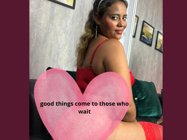NatachaLatinHot with curly brown hair, wearing red lingerie, sitting on a black couch, smiling, showing her back and red lace thigh-highs, with a pink heart covering her ass.