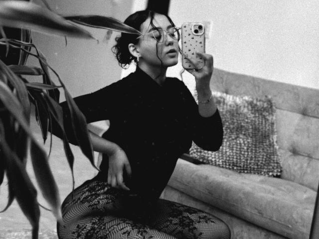 Black and white photo of SophiaBrowm kneeling in front of a mirror. She's wearing a black turtleneck and patterned tights. Her curly hair is up, and she's got glasses on. She's holding a phone with a polka dot case, taking a selfie. A plant is on the left, and a tufted sofa with a textured pillow is in the background. The image has a grainy texture. SophiaBrowm's right hand is on her knee, and her left arm is raised, holding the phone. She looks confident.