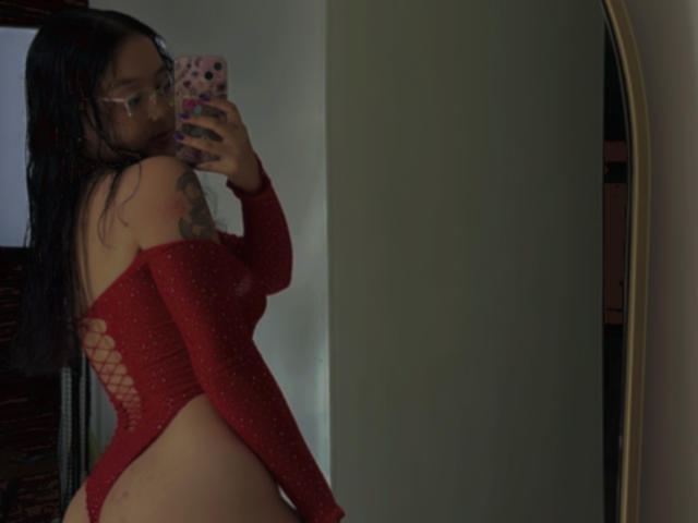 SophiaBrowm with curly black hair in a ponytail, wearing a backless, beige, thong dress, holding a phone, showing her ass, in a room with a balcony.