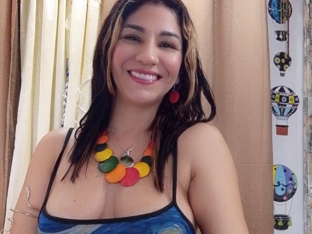 DeliaPin with medium tits, smiling, wearing a blue patterned top, colorful necklace, red earrings, and brown wavy hair, standing in front of beige curtains.