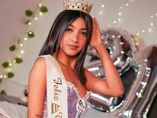 Scarlesth with long black hair, crown, and sash, showing off her tits, hand on head, standing with shiny silver balloons in the background. Warm, soft lighting.