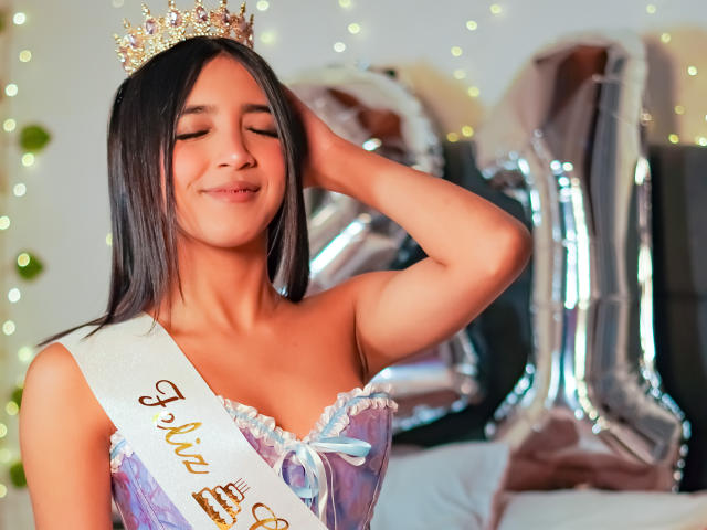 Scarlesth with eyes closed, smiling, wearing a sparkly crown, sash over tits, purple dress with white frills, hand in hair, silver balloons in background.