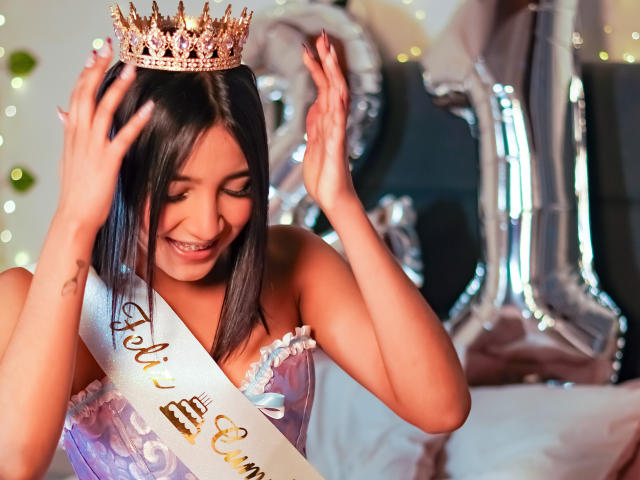 Scarlesth, met een medium bruine huid, zwart steil haar, draagt een roze en witte kroon, glimlachend, handen omhoog, een paars en witte sjerp, zilveren ballonnen, zittend op een bed, glimlachend..