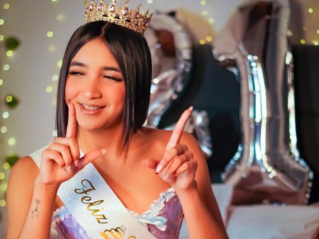 Scarlesth, avec ses courts cheveux noirs, portant une couronne dorée, souriante et pointant du doigt près de ses lèvres. Elle porte une robe violette, mettant en valeur sa poitrine..