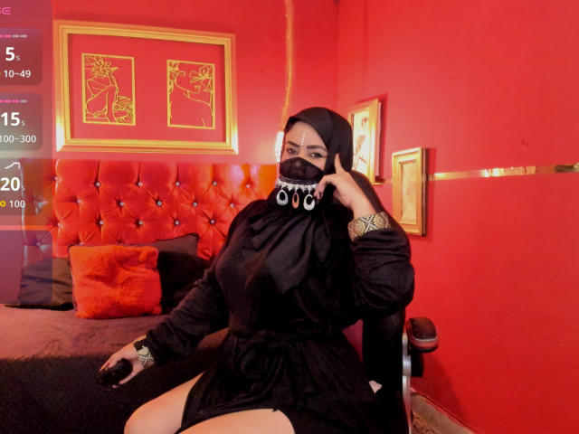 Anissa Ali, une femme aux formes généreuses avec de grandes seins, portant un masque noir, une robe et un foulard, est assise sur une chaise dans une pièce rouge avec des accents dorés et des coussins orange..